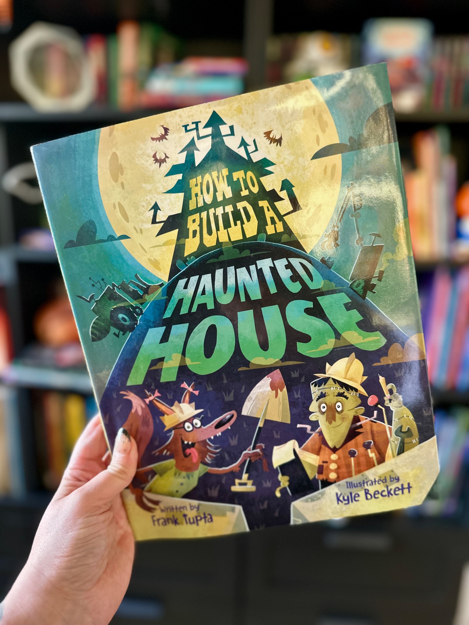 A hand with star-decorated nails holds How to Build a Haunted House before softly blurred library shelves; photograph by October Jones