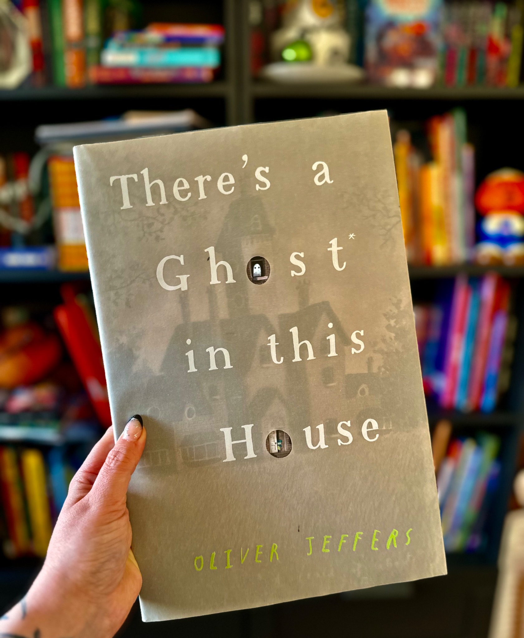 A hand with star-decorated nails holds There's a Ghost in This House before softly blurred library shelves; photograph by October Jones