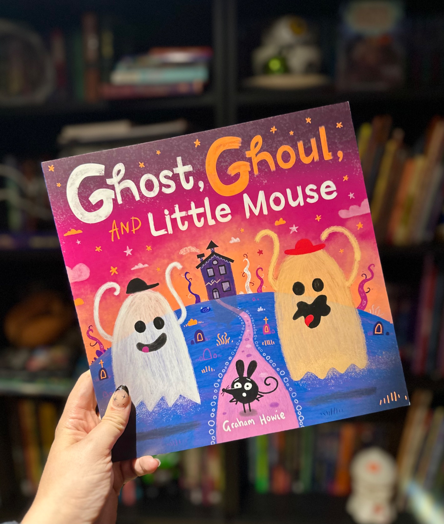 A hand holds Ghost, Ghoul, and Little Mouse in front of softly blurred library shelves