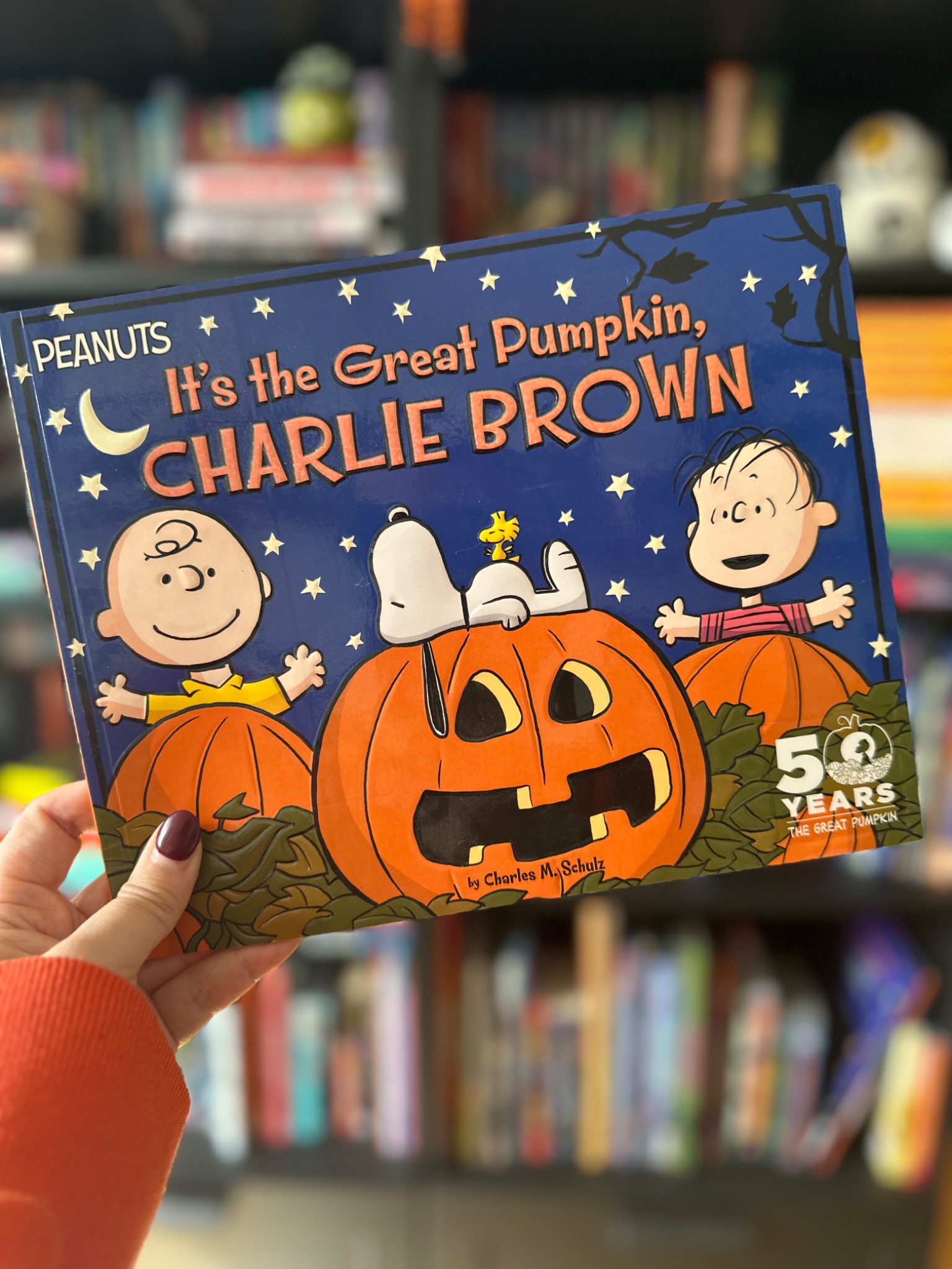 A hand in an orange sleeve holds It's the Great Pumpkin, Charlie Brown in front of softly blurred library shelves