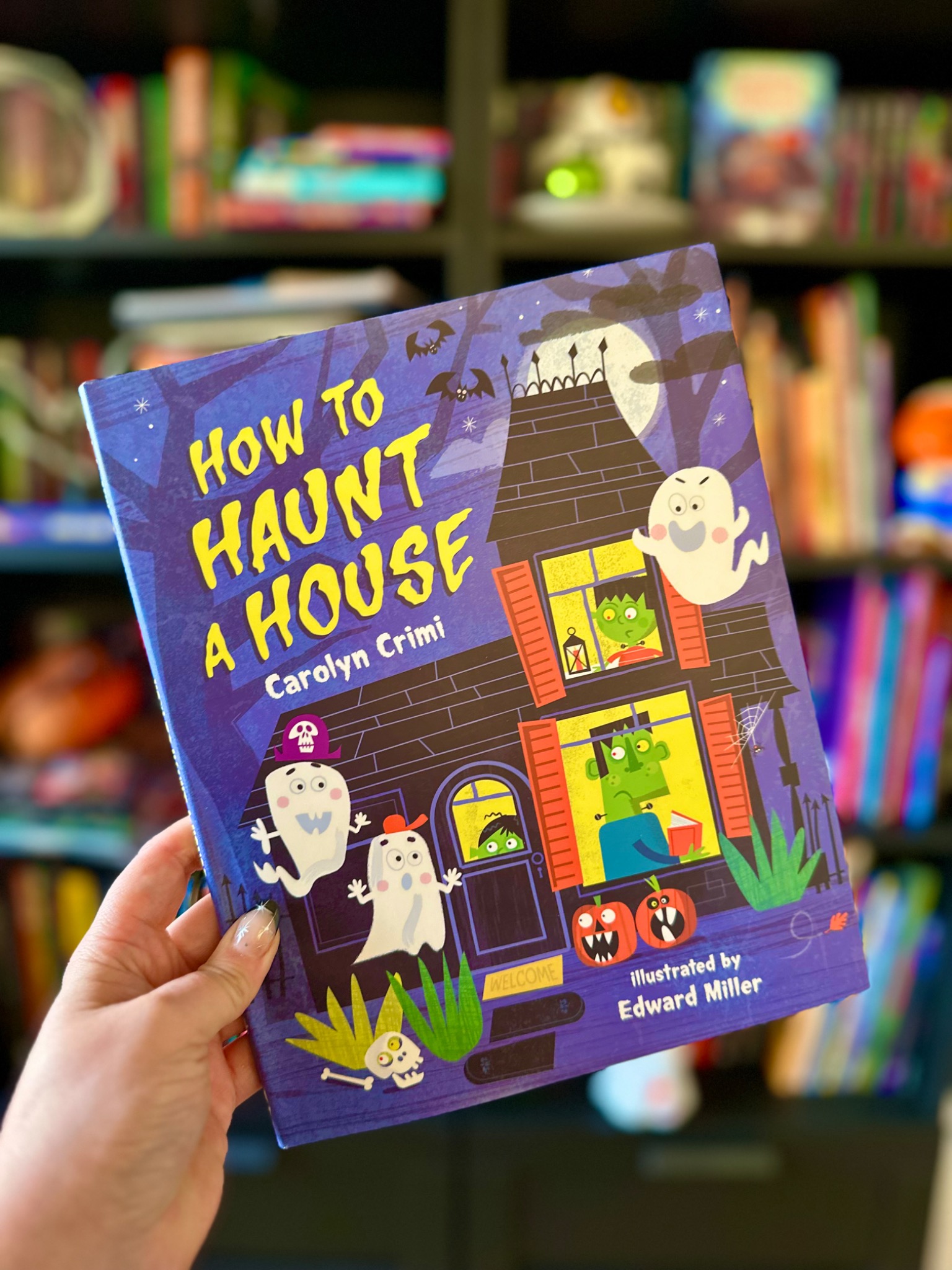 A hand with star-decorated nails holds How to Haunt a House before softly blurred library shelves; photograph by October Jones