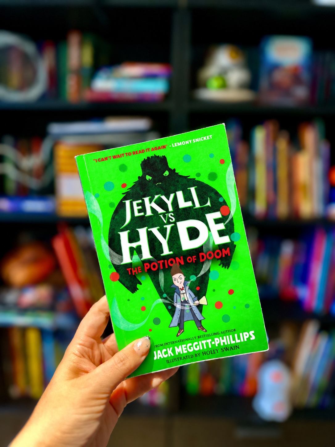 A hand with star-decorated nails holds Jekyll vs Hyde: The Potion of Doom before blurred library shelves; the published bright-green cover has been composited onto October Jones’s proof-copy photograph