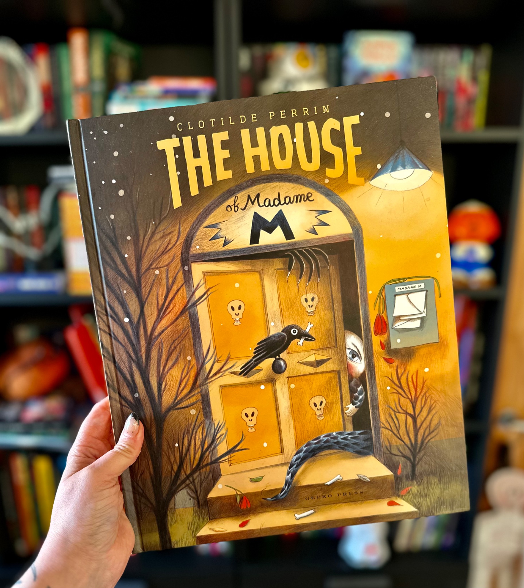 A hand with star-decorated nails holds The House of Madame M before softly blurred library shelves; photograph by October Jones