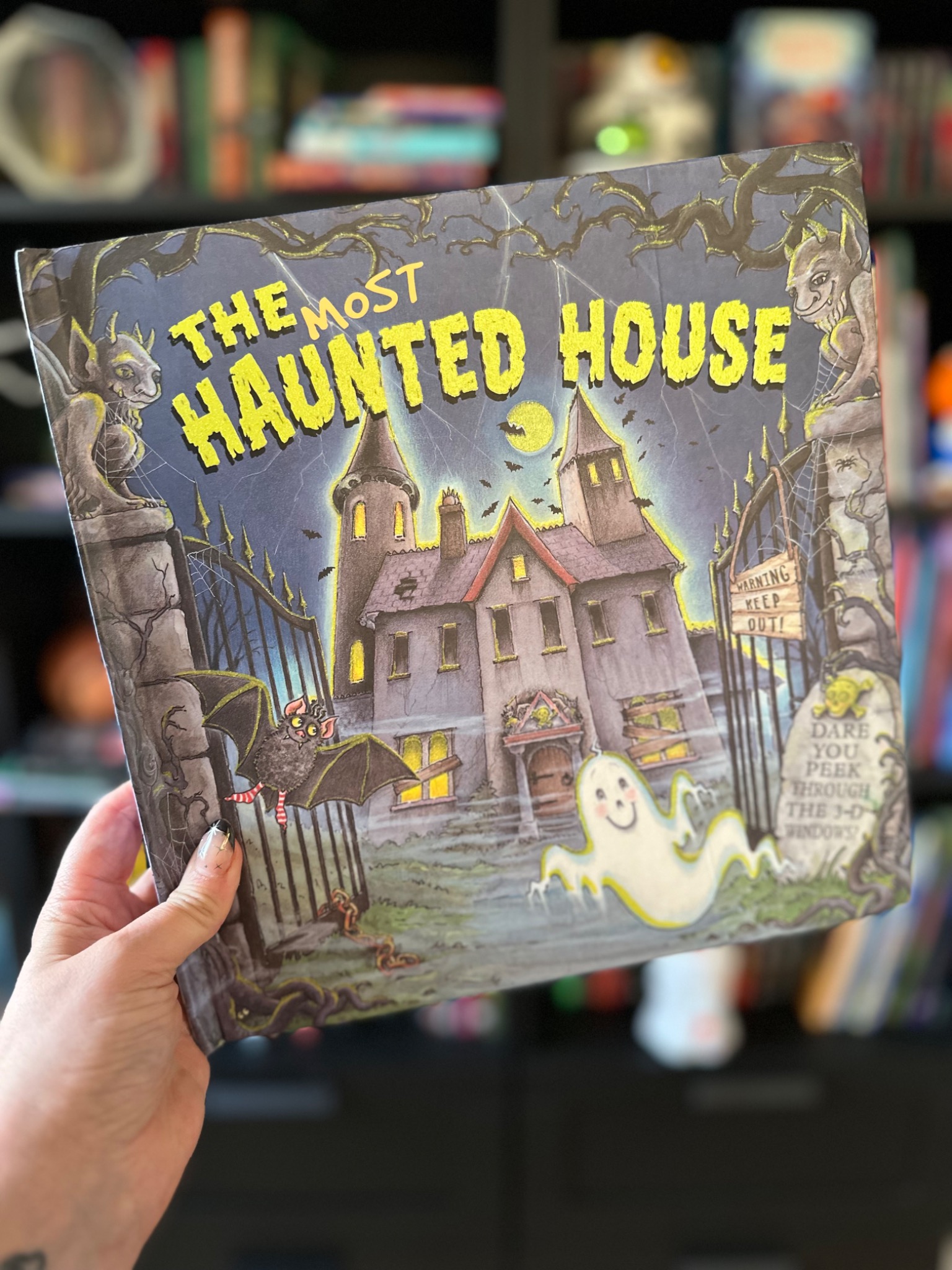 A hand with star-decorated nails holds The Most Haunted House before softly blurred library shelves; photograph by October Jones