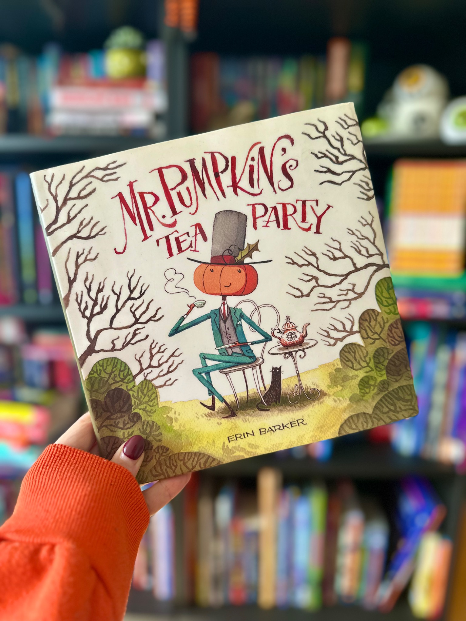 A hand in an orange sleeve holds Mr. Pumpkin's Tea Party in front of softly blurred library shelves