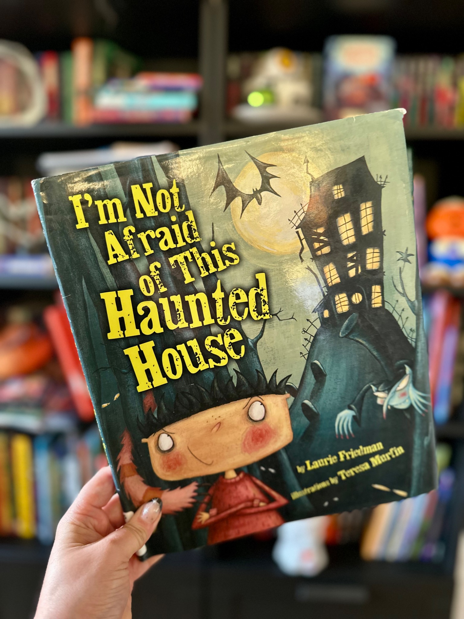 A hand with star-decorated nails holds I'm Not Afraid of This Haunted House before softly blurred library shelves; photograph by October Jones