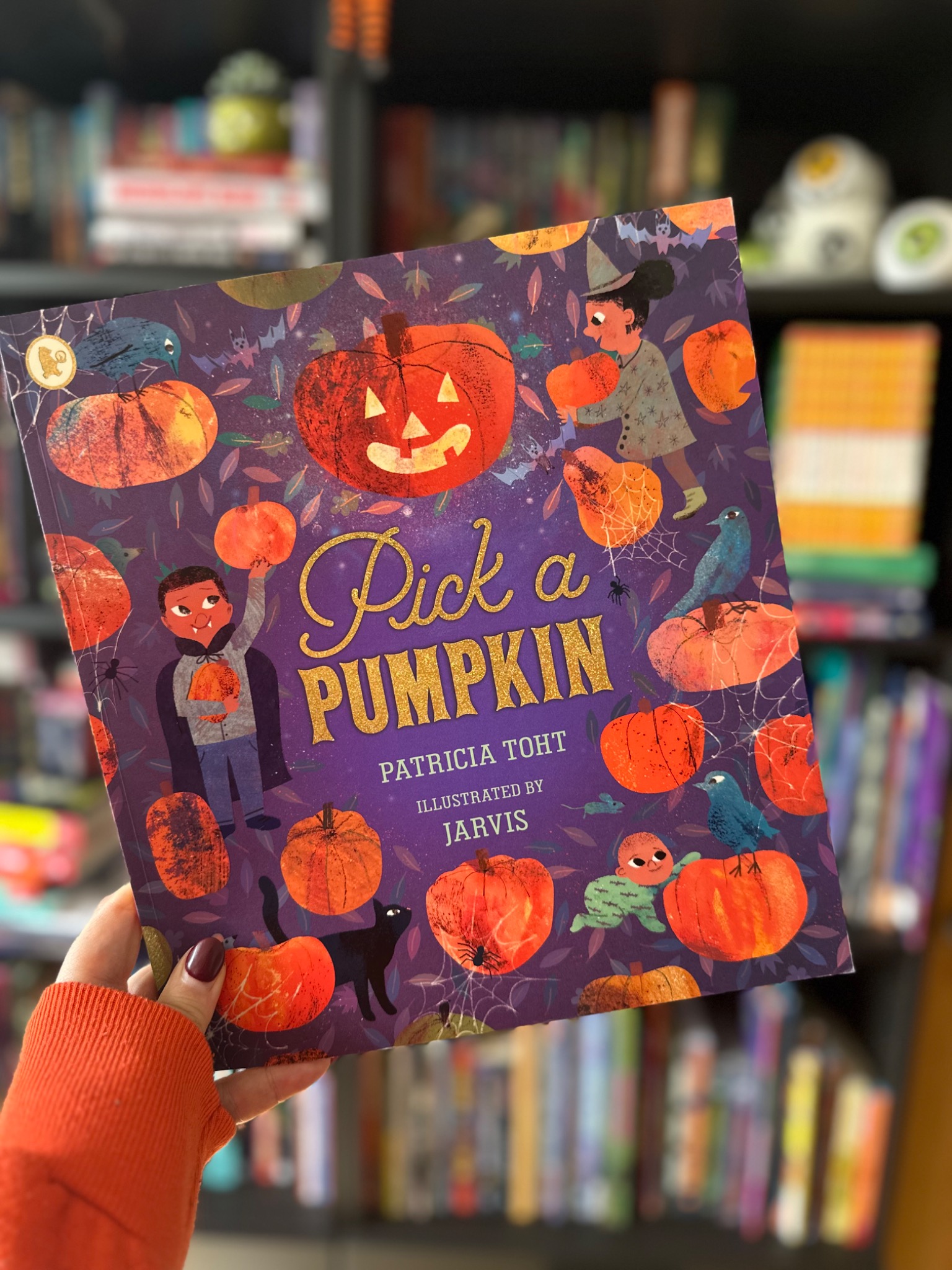 A hand in an orange sleeve holds Pick a Pumpkin in front of softly blurred library shelves