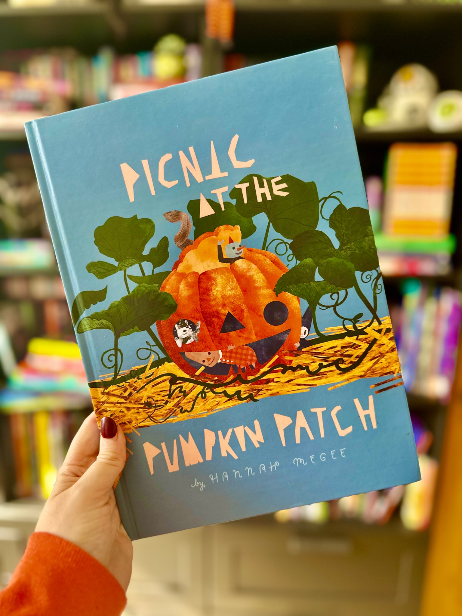 A hand holds Picnic at the Pumpkin Patch by Hannah Megee in front of softly blurred library shelves