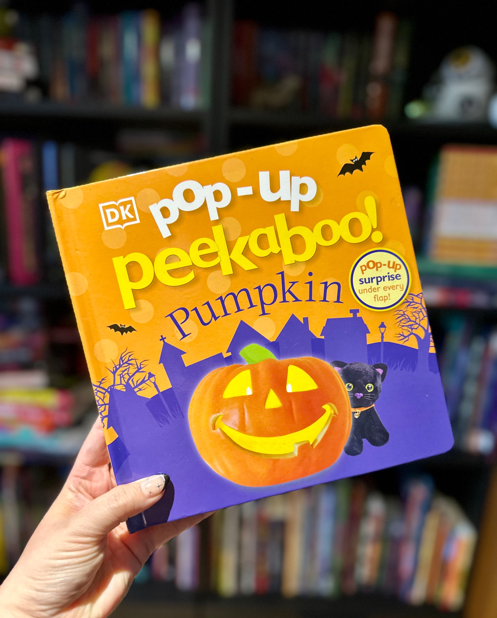 A hand holds the orange and purple Pop-Up Peekaboo! Pumpkin book in front of softly blurred library shelves