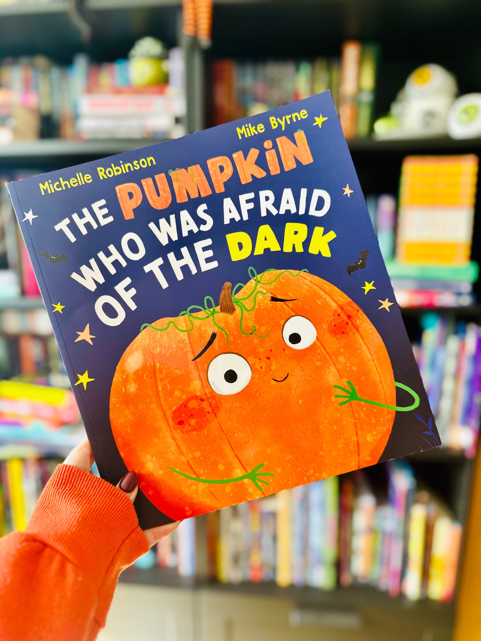 A hand in an orange sleeve holds The Pumpkin Who Was Afraid of the Dark by Michelle Robinson and Mike Byrne in front of blurred bookshelves