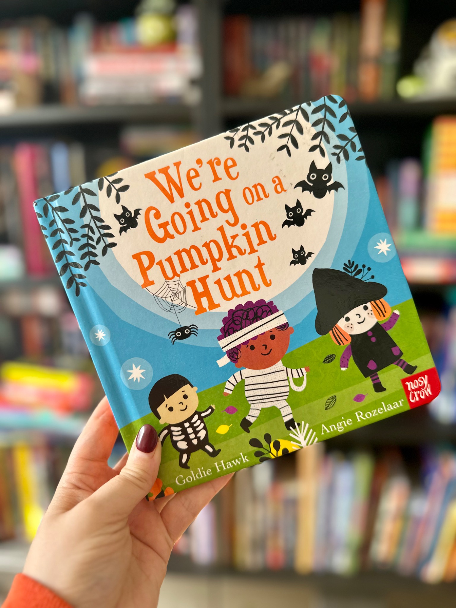 A hand in an orange sleeve holds We're Going on a Pumpkin Hunt! in front of softly blurred library shelves