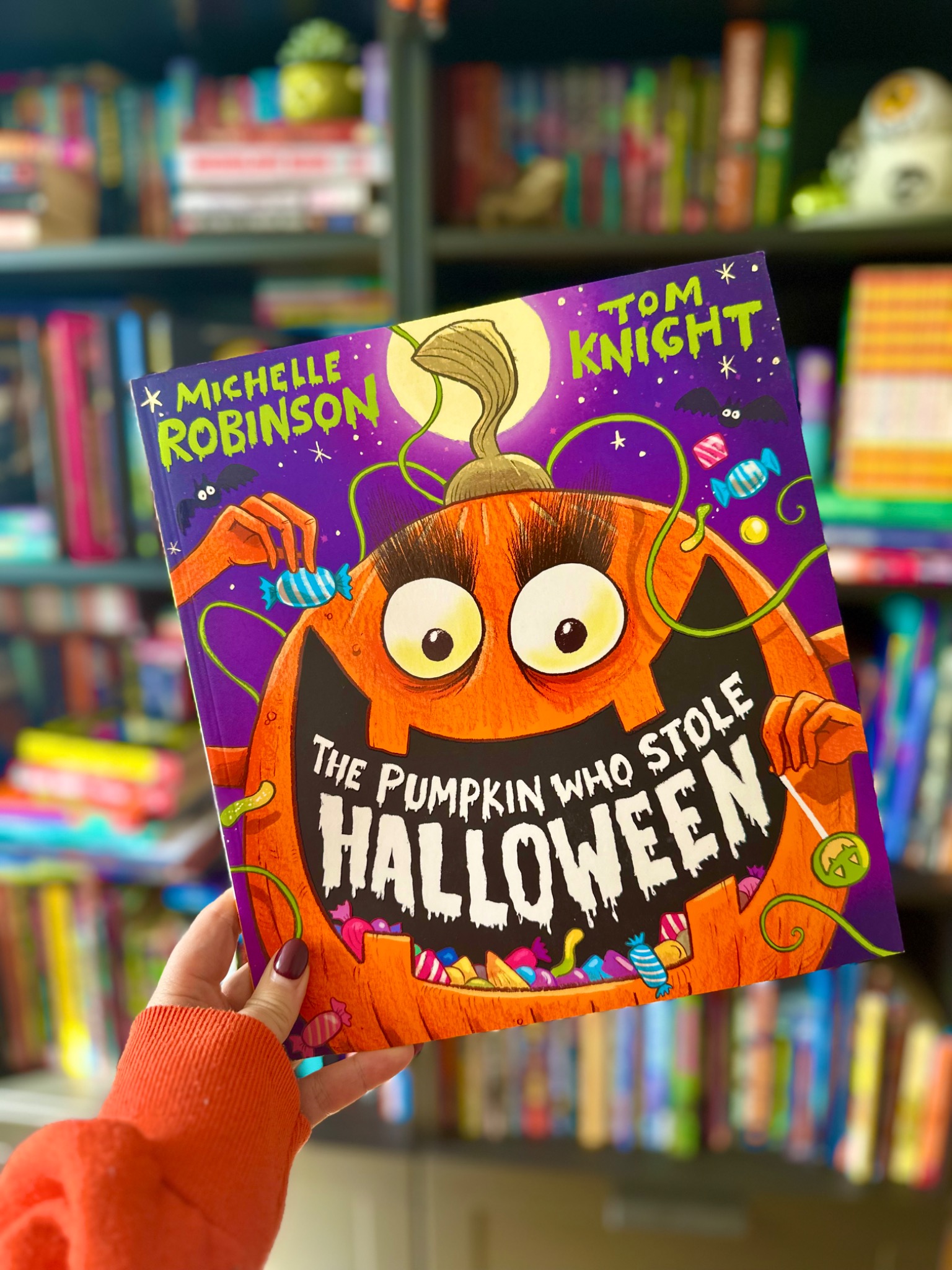 A hand in an orange sleeve holds The Pumpkin Who Stole Halloween in front of softly blurred library shelves
