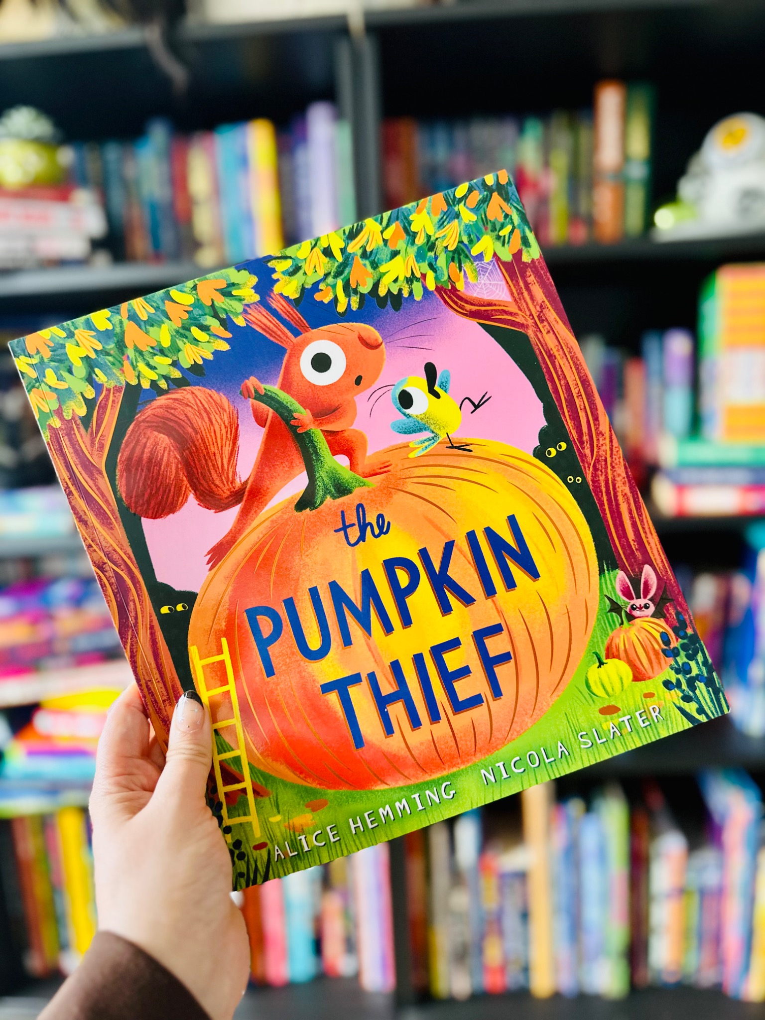A hand holds The Pumpkin Thief in front of softly blurred library shelves