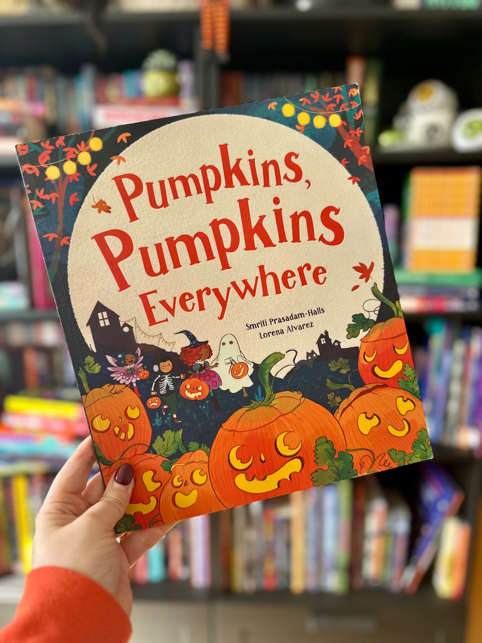 A hand in an orange sleeve holds Pumpkins, Pumpkins Everywhere in front of softly blurred library shelves