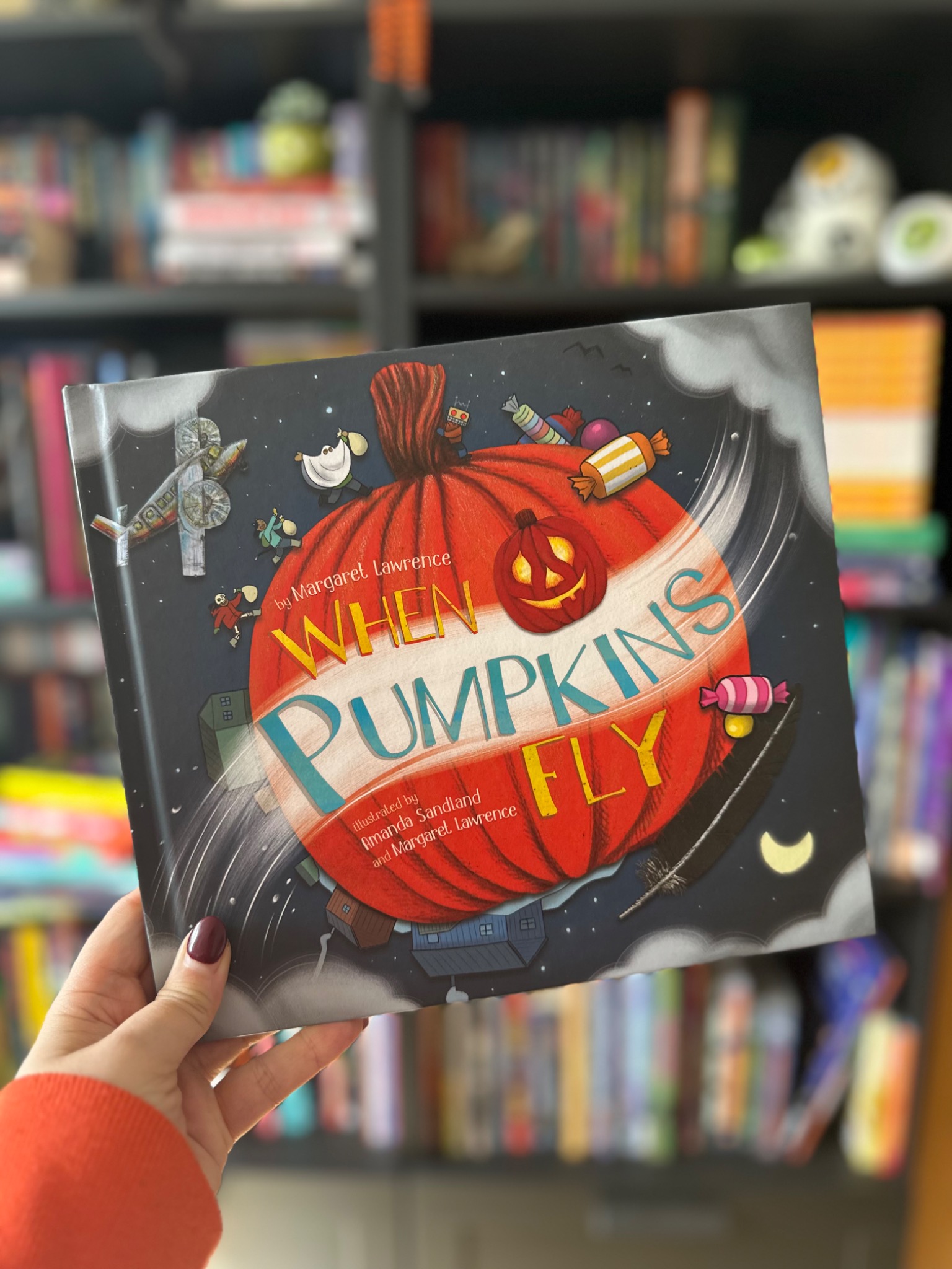 A hand in an orange sleeve holds When Pumpkins Fly in front of softly blurred library shelves