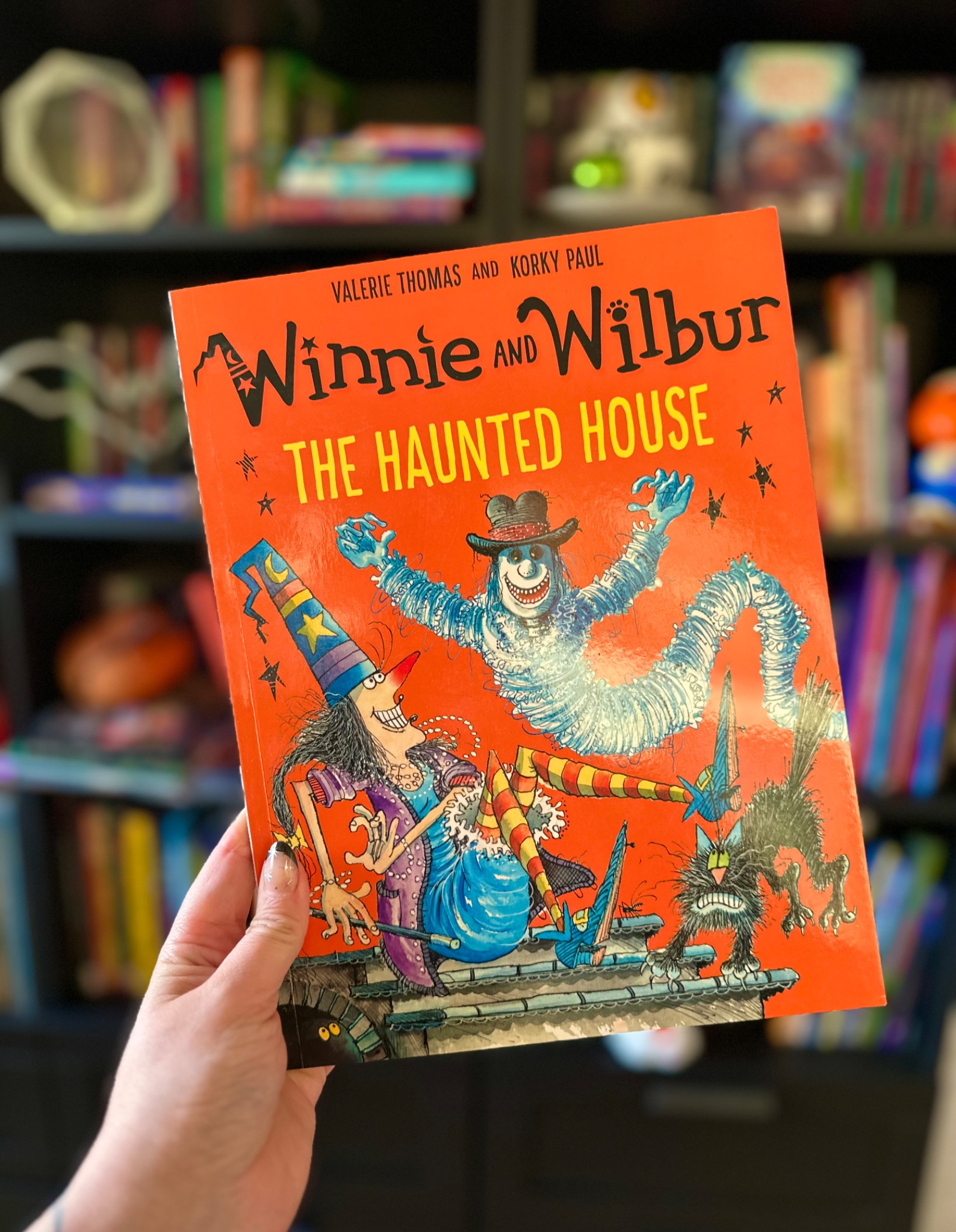 A hand with star-decorated nails holds Winnie and Wilbur: The Haunted House before softly blurred library shelves; photograph by October Jones