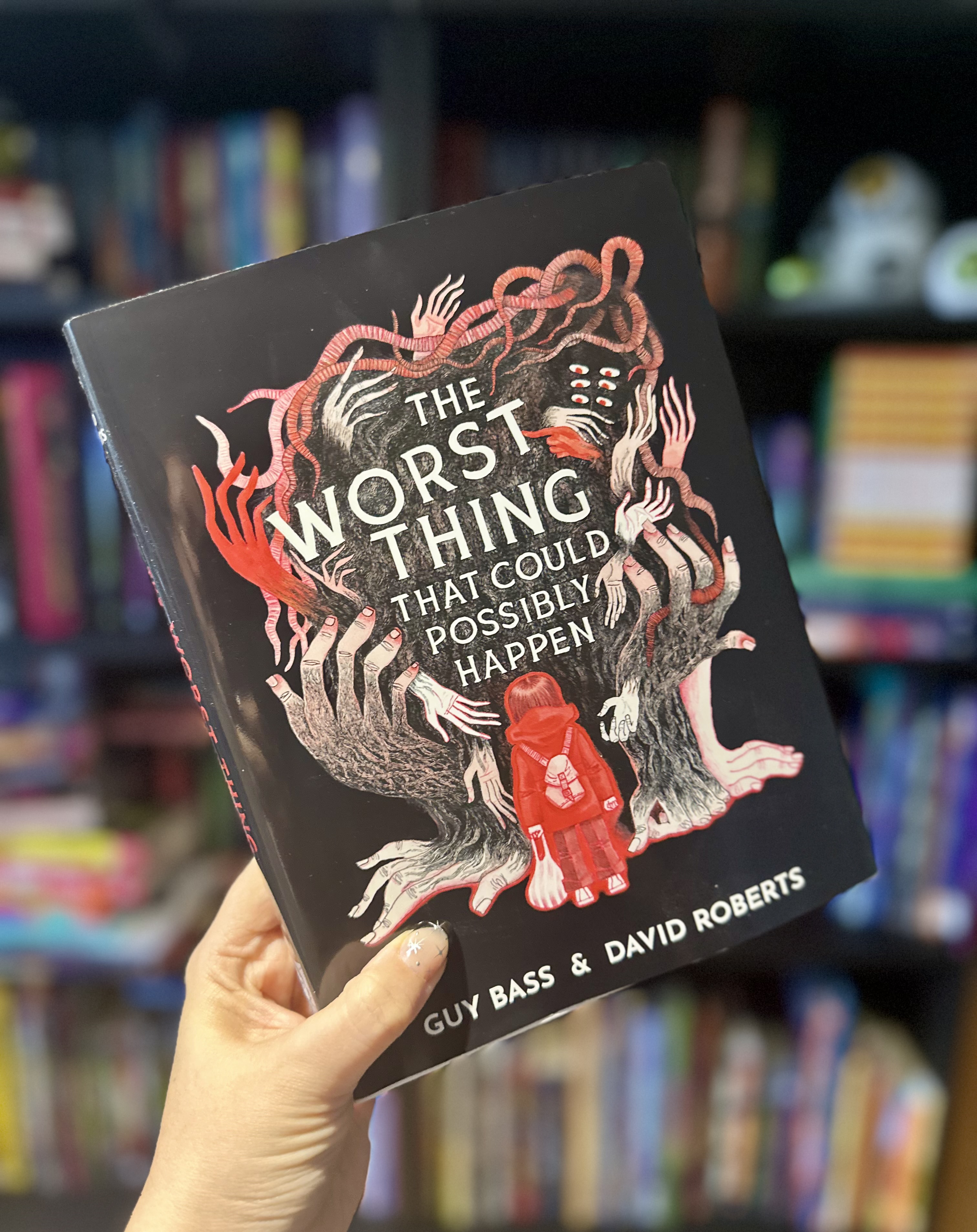 A hand holds The Worst Thing That Could Possibly Happen in front of softly blurred bookshelves